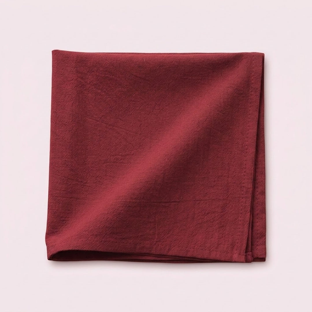 A folded square piece of textured fabric in a deep burgundy red, displayed on a solid pale pink background.