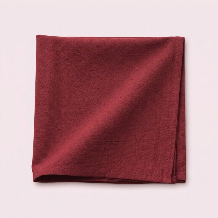 A folded square piece of textured fabric in a deep burgundy red, displayed on a solid pale pink background.