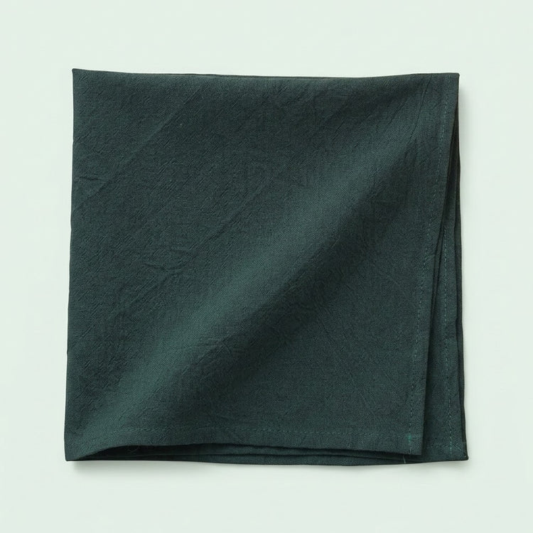 A folded dark forest green fabric napkin or pocket square with a subtle crinkled texture, displayed on a light off-white background.