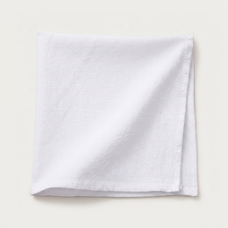 A folded white cotton napkin with a textured weave, displayed on a light beige background.