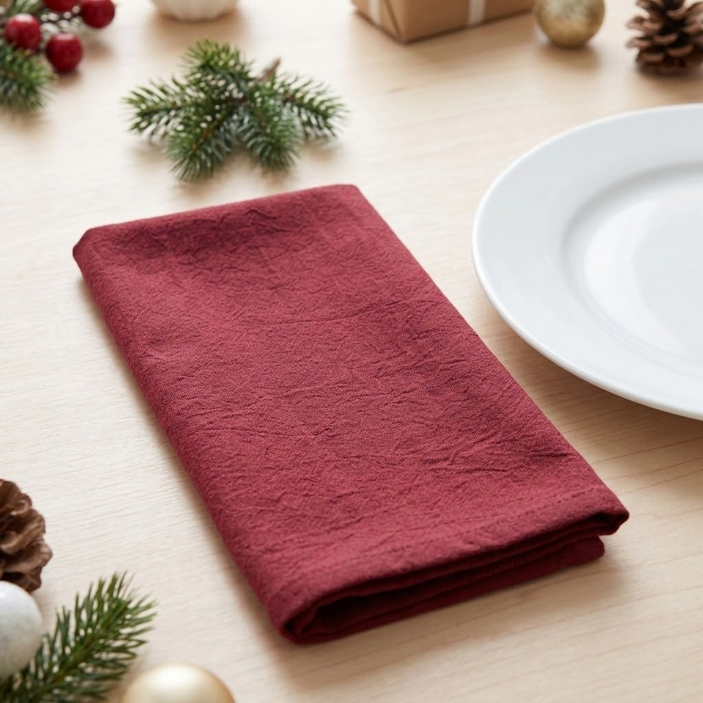 A folded dark red linen napkin with a crinkled texture, styled on a light wood table with holiday decorations like pine branches, ornaments, and a white plate.
