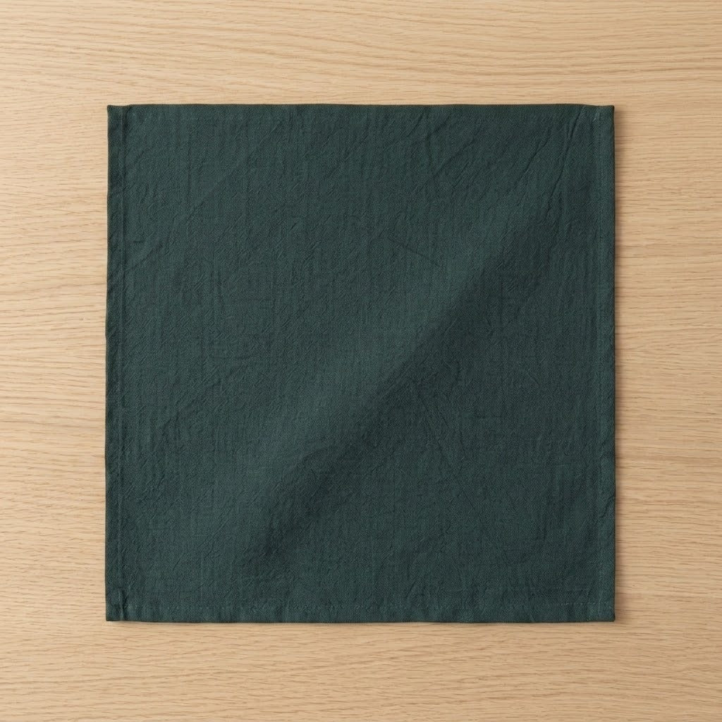 A square dark green fabric napkin with a crinkled texture, laid flat on a light wood grain surface.