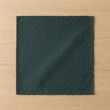 A square dark green fabric napkin with a crinkled texture, laid flat on a light wood grain surface.