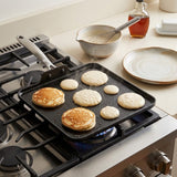 A black non-stick square griddle pan with a white handle cooking pancakes on a gas stovetop in a kitchen setting.