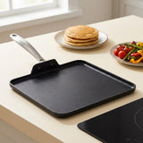 A square black non-stick griddle pan with a silver metal handle, shown on a light beige countertop next to an induction cooktop, with pancakes and a salad in the background.