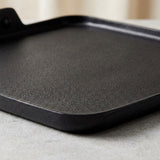 A close-up of a black cast iron griddle or flat pan with a textured surface and a handle visible in the upper left, resting on a light gray surface.