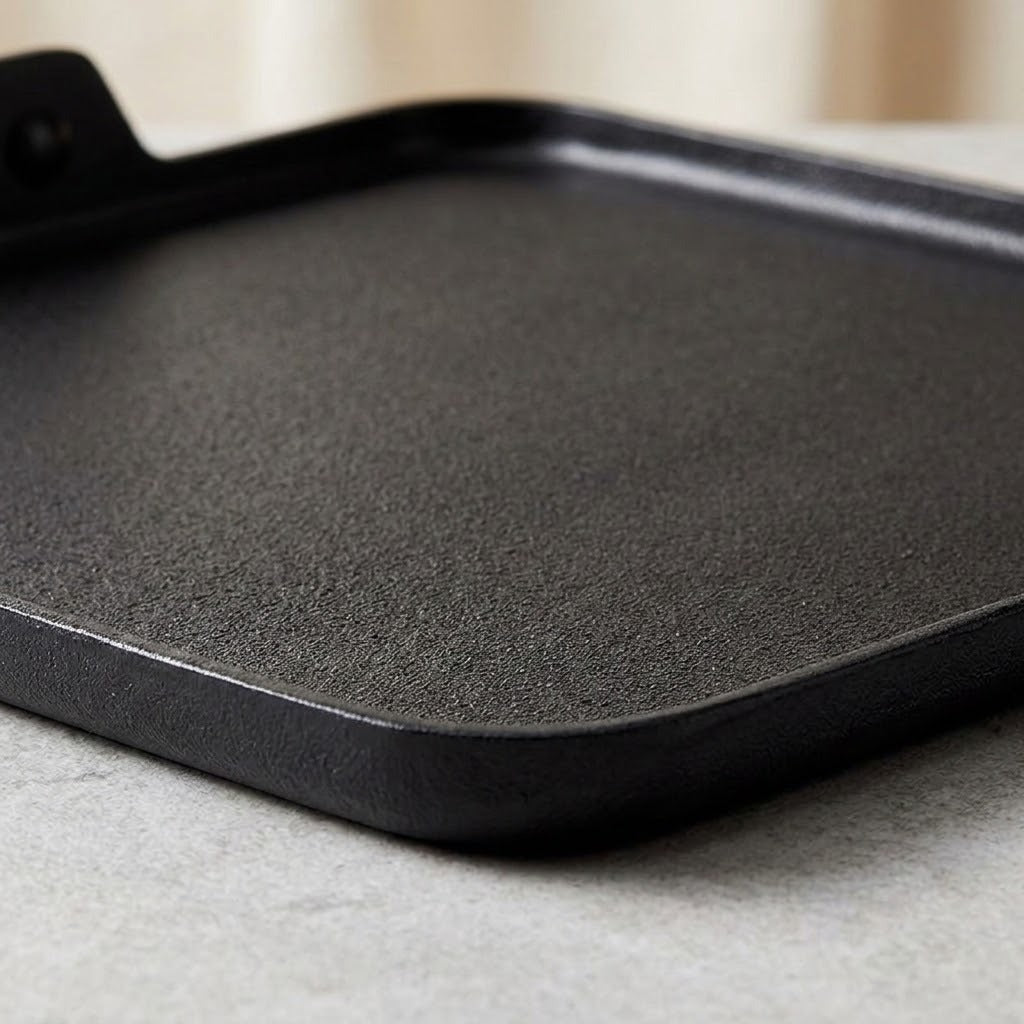 A close-up of a black cast iron griddle or flat pan with a textured surface and a handle visible in the upper left, resting on a light gray surface.