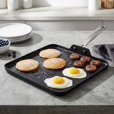A black square griddle pan with a silver handle sits on a gray kitchen countertop, cooking pancakes, eggs, sausage, and blueberries. White plates and a bowl of blueberries are visible in the softly blurred background kitchen setting.