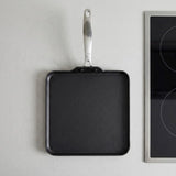 A square black non-stick griddle pan with a silver metal handle, shown hanging against a light gray wall next to a black induction cooktop.