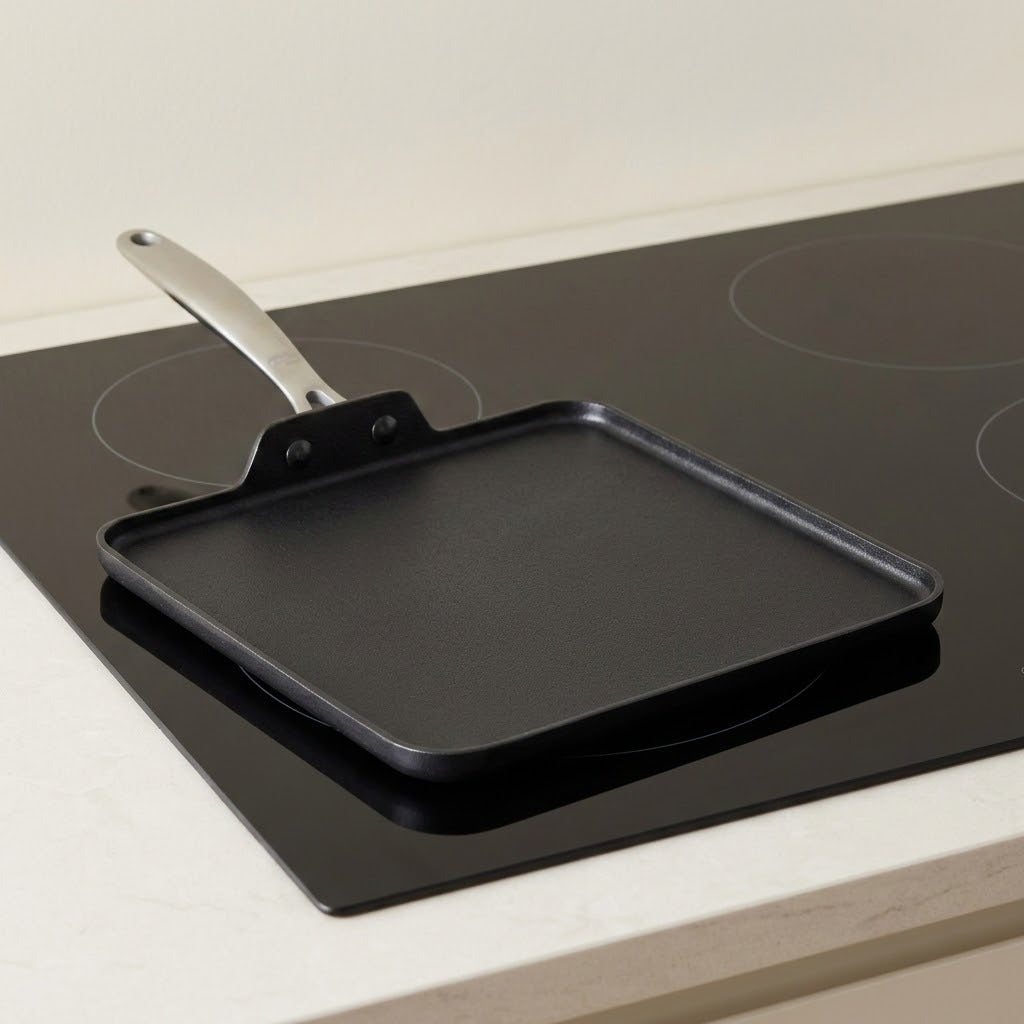 A square black non-stick griddle pan with a silver metal handle, shown resting on a black electric stovetop cooktop.