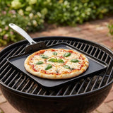 A square black cast-iron skillet with a handle sits on the grill grate of a round charcoal kettle, cooking a small pizza topped with melted cheese and fresh basil leaves.