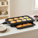 A black multi-cooker electric griddle on a white countertop, cooking pancakes and two sunny-side-up eggs. A tray underneath holds cooked pancakes, and the background shows a softly blurred kitchen setting with shelves, bowls, and a bowl of berries.