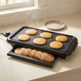Black electric griddle with pancakes cooking on the flat top and a tray holding more pancakes below, set on a kitchen counter near a window.