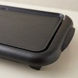 Close-up of a black electric griddle or flat-top pan showing the matte cooking surface and side handle on a light neutral background.