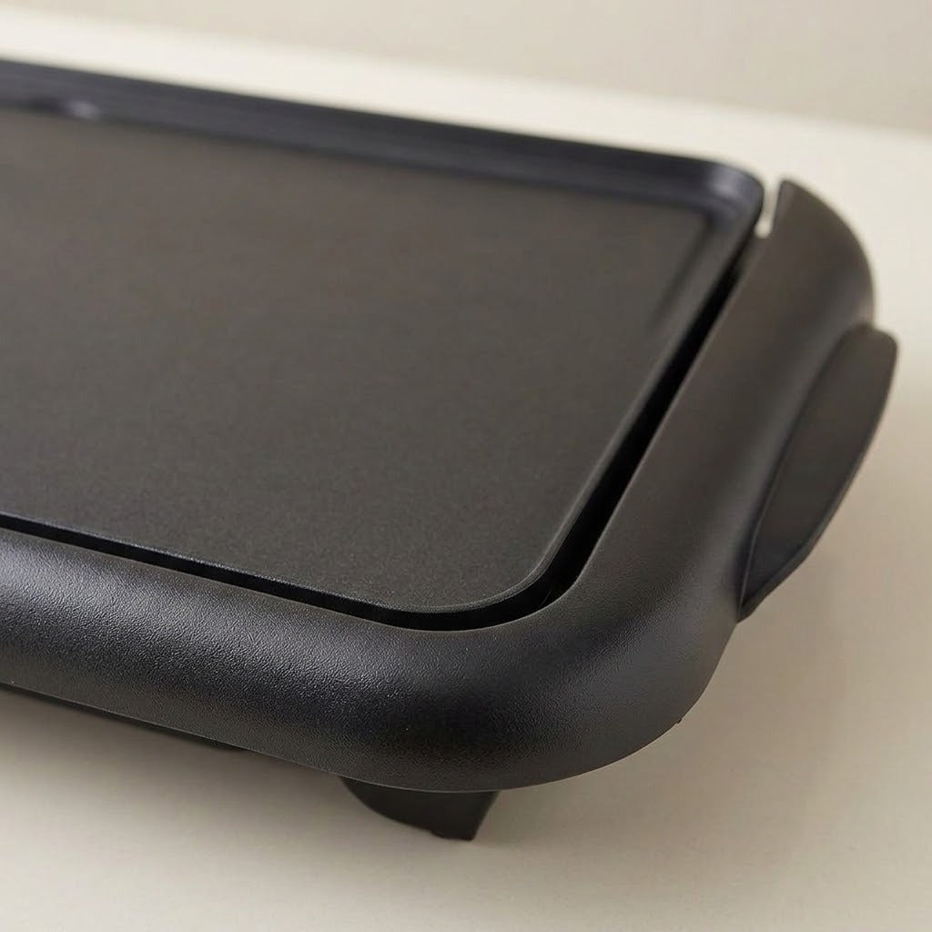 Close-up of a black electric griddle or flat-top pan showing the matte cooking surface and side handle on a light neutral background.