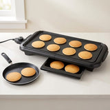 Black electric mini pancake maker with multiple cooking plates, shown on a white countertop with several golden pancakes cooking inside. A small detachable frying pan is placed beside it.