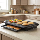 A black electric griddle with a lower tray sits on a kitchen counter, cooking pancakes and vegetables. The background shows a blurred kitchen interior with beige cabinets and a tiled backsplash.