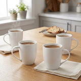 Four white ceramic mugs filled with coffee arranged on a light wood table in a bright kitchen setting, with napkins and a plate of toast nearby.