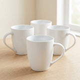Four white ceramic mugs arranged on a light wood tabletop near a bright window.