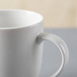 A close-up of a glossy white ceramic mug with a thick handle, set against a softly blurred background with a gray lower section and a light beige upper section.
