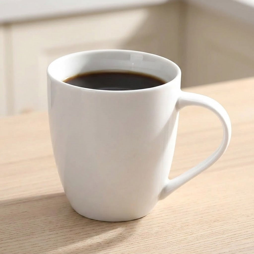 A white ceramic coffee mug filled with black coffee, sitting on a light wood table in a softly lit indoor setting.
