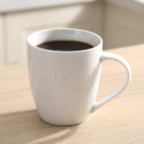 A white ceramic coffee mug filled with black coffee, sitting on a light wood table in a softly lit indoor setting.