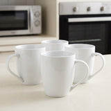 Four white ceramic mugs arranged on a light countertop in a kitchen setting, with a blurred microwave and oven in the background.