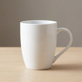 A plain white ceramic mug with a curved handle, sitting on a light wood tabletop against a neutral gray background.