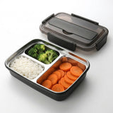 A black and stainless steel compartment lunch box with food inside, shown open with its lid placed behind it on a light gray background.