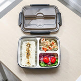 A stainless steel 3-compartment bento lunch box with a clear plastic lid, shown open on a light wood table. The compartments are filled with white rice, spring roll wrappers, and a small salad with red tomatoes.