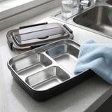 A stainless steel three-compartment lunch tray with a black outer base and a translucent lid, shown on a kitchen countertop next to a sink with a blue towel draped over the side.