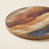 A round wooden coastert or trivet with a natural wood grain pattern in shades of brown, beige, and gray, shown on a light cream background.