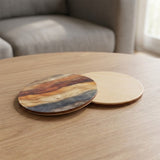 Two round wooden coasters on a light wood table in a living room setting, with one coaster showing a multicolor natural wood grain pattern and the other showing a smooth light natural wood finish.