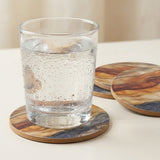 A clear glass tumbler with condensation sits on a round wood coaster, with two additional matching coasters behind it on a light tabletop.