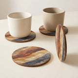 Three round wooden coasters with a wavy multicolor grain pattern in brown, beige, and blue, shown with two matching ceramic cups on a light cream background.