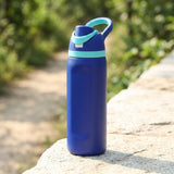 A blue water bottle with a mint green lid and carrying loop, standing on a stone ledge with a blurred outdoor background.