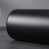 A close-up of a matte black cylindrical product, possibly a cup, tumbler, or container, resting on a dark gray surface against a neutral gray background.
