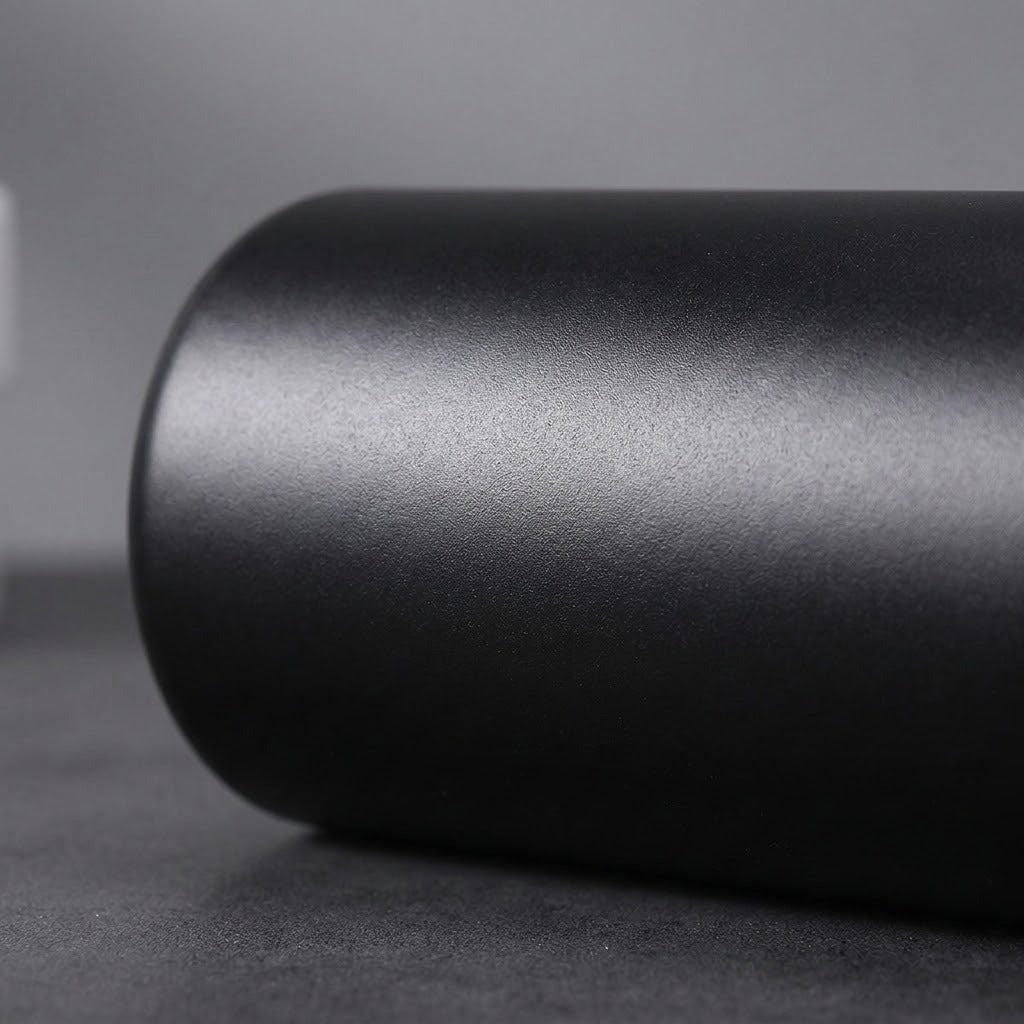A close-up of a matte black cylindrical product, possibly a cup, tumbler, or container, resting on a dark gray surface against a neutral gray background.