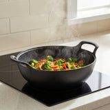A black cast iron wok with handles sits on a black induction cooktop in a kitchen, cooking a steaming mixture of broccoli, carrots, and red peppers.