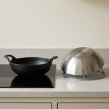 A black cast iron kadhai with side handles sits on a black induction cooktop, next to a stainless steel covered dish on a marble kitchen countertop.