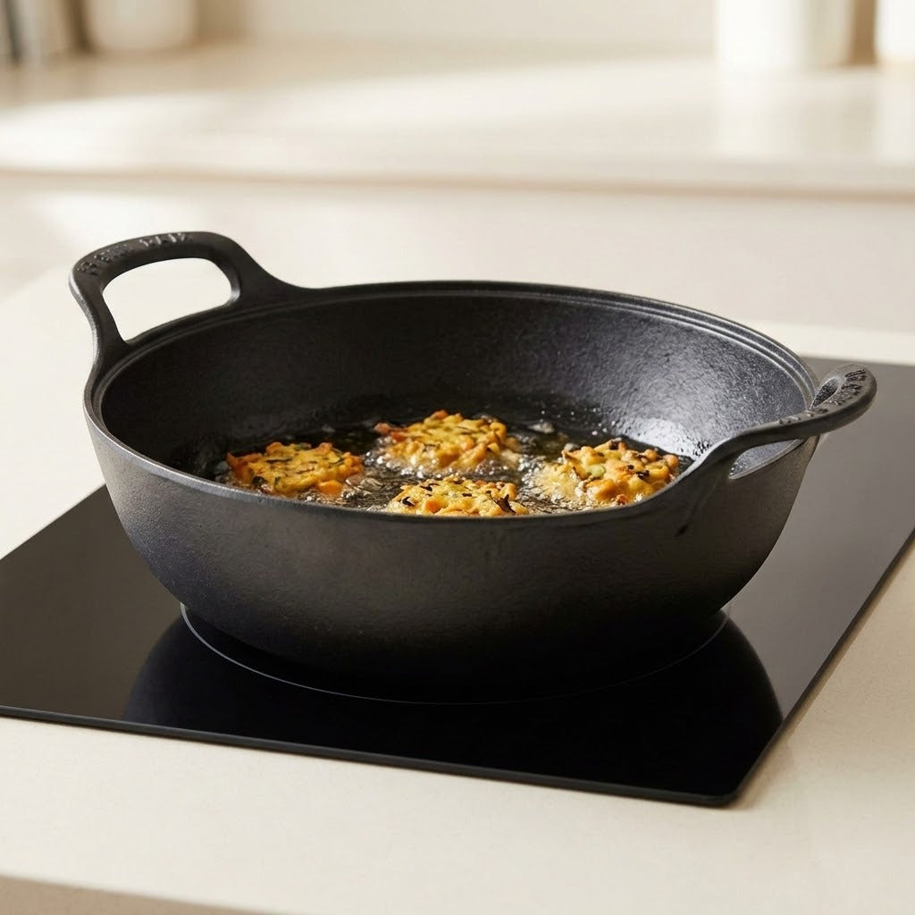 A black cast iron skillet with two handles sits on a black induction cooktop, frying golden-brown food in oil. The background is a softly blurred kitchen setting.