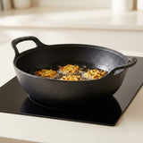 A black cast iron skillet with two handles sits on a black induction cooktop, frying golden-brown food in oil. The background is a softly blurred kitchen setting.