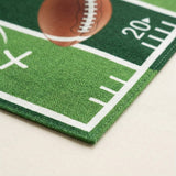 A close-up of a green football field printed rug or mat with white yard line markings, a brown football graphic, and the number 20, shown on a light beige surface.
