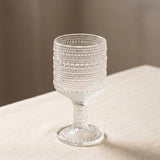 A clear textured glass goblet with a beaded bubble pattern, standing on a beige fabric surface against a soft neutral background.