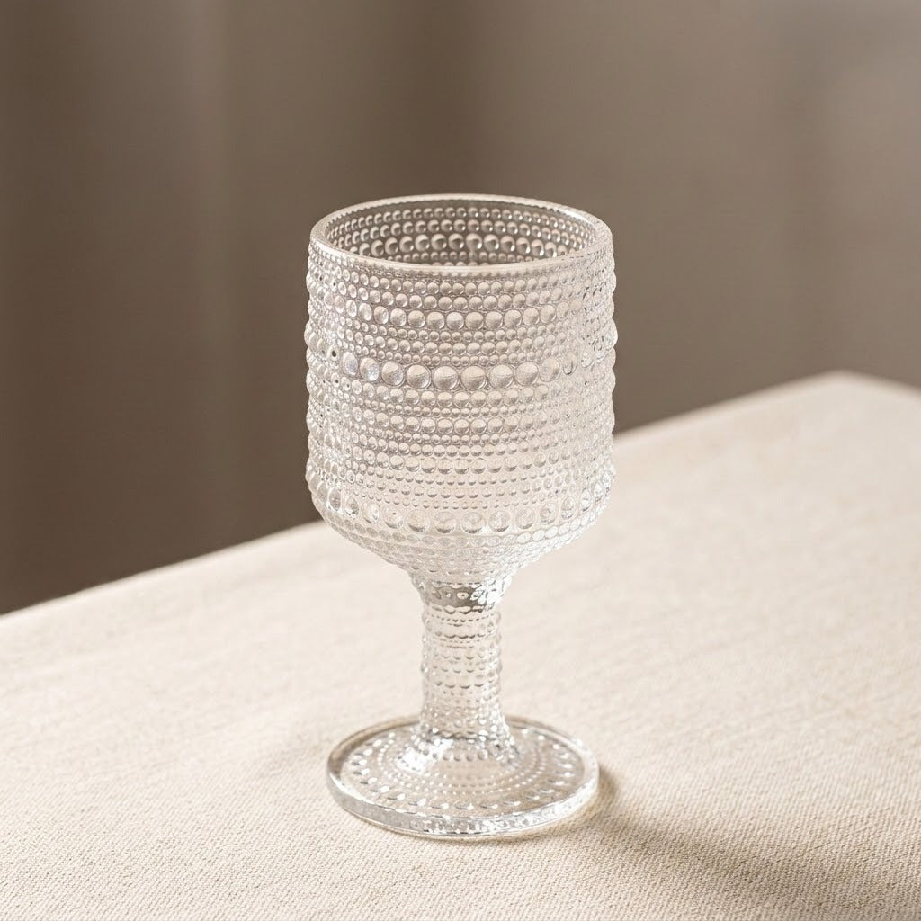 A clear textured glass goblet with a beaded bubble pattern, standing on a beige fabric surface against a soft neutral background.