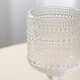 A close-up of a clear glass goblet with a textured, bubble-patterned surface, resting on a light beige surface with a soft gray background.