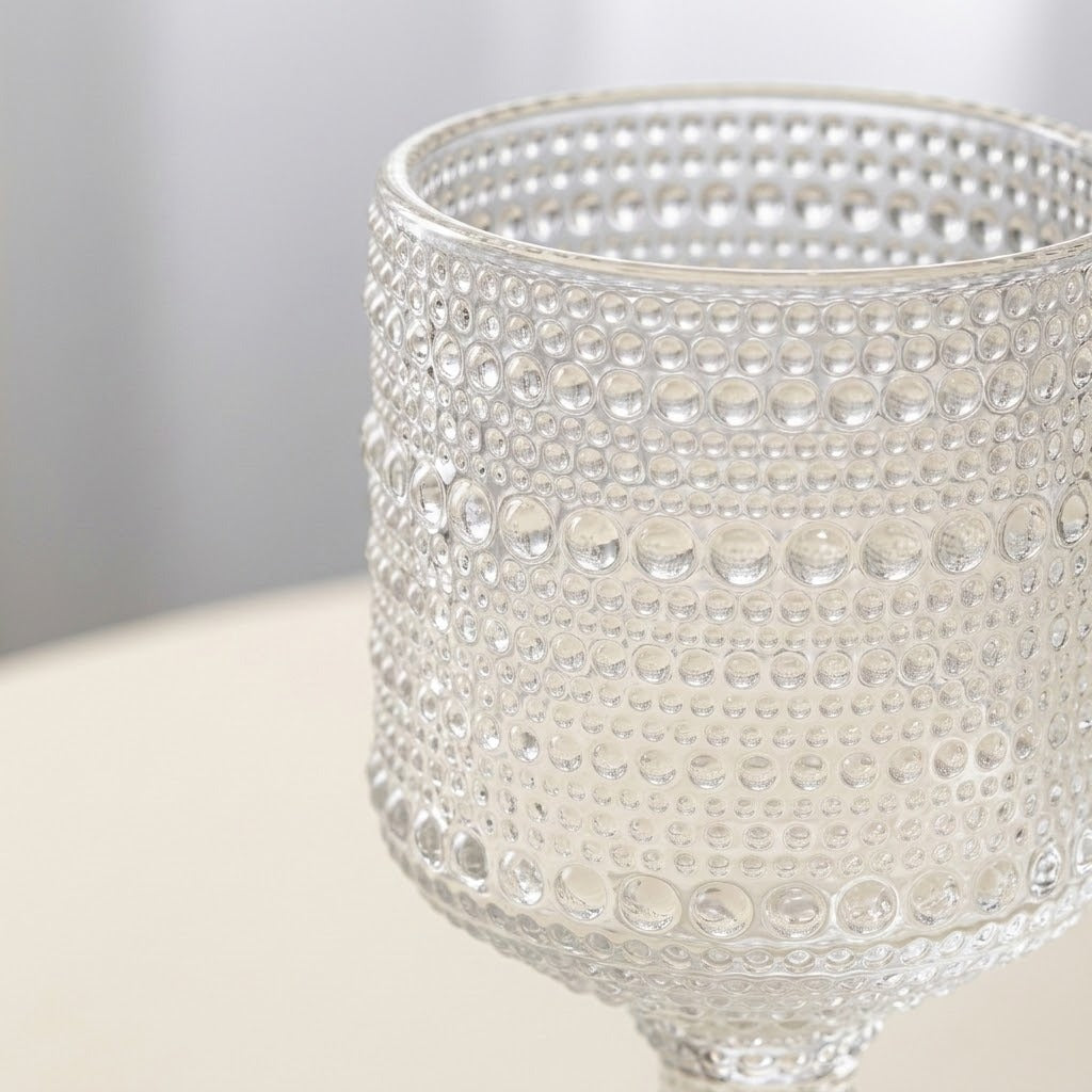A close-up of a clear glass goblet with a textured, bubble-patterned surface, resting on a light beige surface with a soft gray background.