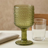 An olive green textured glass goblet with a bubble pattern and a matching stem, shown next to a clear glass tumbler on a tabletop with a neutral background.