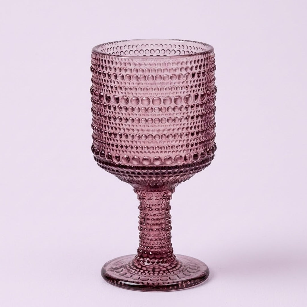 A single textured glass goblet in a dusty rose or mauve purple color, featuring a beaded bubble pattern on the bowl and stem, set against a solid light lavender background.