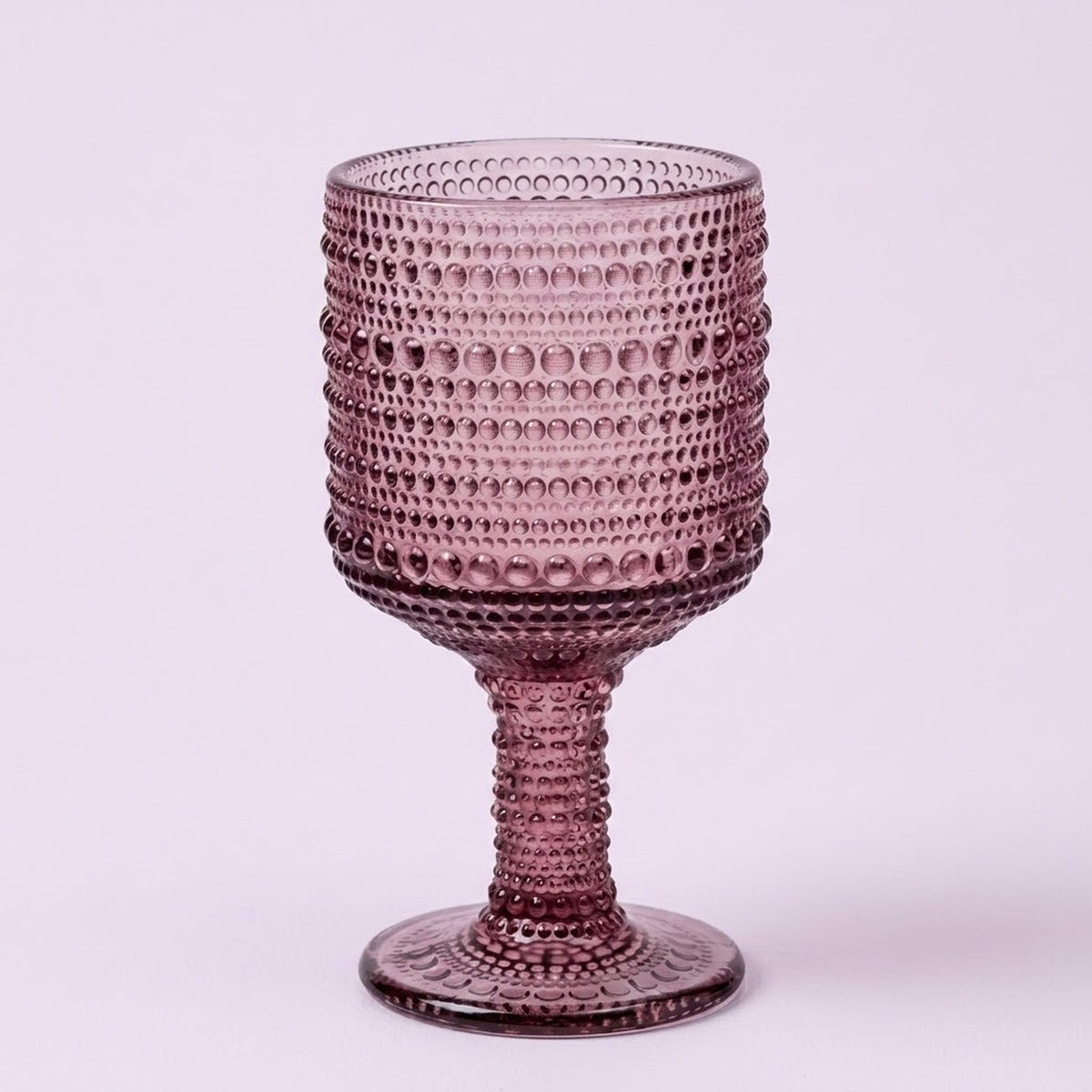 A single textured glass goblet in a dusty rose or mauve purple color, featuring a beaded bubble pattern on the bowl and stem, set against a solid light lavender background.